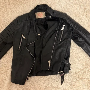 Svart skinnjacka från Voyelles - Snygg svart skinnjacka från Voyelles med coola dragkedjedetaljer och bälte i midjan. Jackan har långa ärmar med quiltade axlar och en asymmetrisk dragkedja framtill. Perfekt för en edgy look!