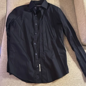 Svart skjorta från Dressmann - Snygg svart skjorta från Dressmann i slim fit. Tillverkad i premium bomullsstretch för extra komfort. Perfekt för en stilren look med knappar framtill och klassisk krage.