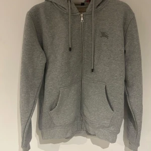 Grå hoodie från Burberry - Säljer en stilren grå hoodie med dragkedja och justerbar huva. Passar som en M.