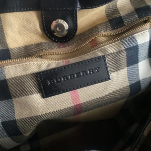 Svart axelväska från Burberry - Snygg svart axelväska från Burberry med klassiskt rutigt innerfoder. Köptes i Hongkong 2013 för 6900 HKD vilket motsvarar ungefär 9750kr  Vintage modellen heter ”trench group small canterbury tote”. De säljs inte längre. 