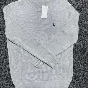 Grå stickad tröja från Polo Ralph Lauren - Snygg grå stickad tröja från Polo Ralph Lauren med det klassiska broderade logotypen i blått på bröstet. Tröjan har en rund halsringning och långa ärmar, perfekt för kyligare dagar. 