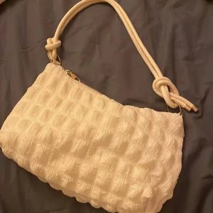 Väska - Snygg vit axelväska med unik knutdetalj på axelremmen. Väskan har en quiltad textur och stängs med dragkedja. ✨