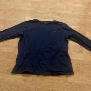 Mörkblå långärmad tröja från Polo Ralph Lauren - Säljer en stilren mörkblå långärmad tröja från Polo Ralph Lauren. Tröjan har en klassisk design med det ikoniska röda logotypbroderiet på bröstet. Perfekt för en avslappnad och snygg look.