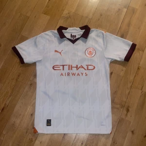 Manchester city tröja 23/24 bortatröja - Säljer en vit fotbollströja från Puma med bruna detaljer och korta ärmar. Tröjan har trycket 'Gvardiol 24' på ryggen och 'Etihad Airways' på framsidan. Helt oanvänd.