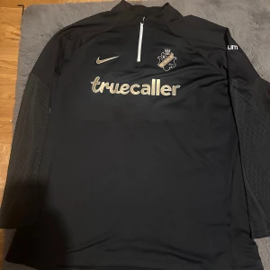 Svart långärmad fotbollströja från Nike - Snygg svart långärmad fotbollströja från Nike med AIK-logga och Truecaller-tryck på framsidan. Tröjan har även Stadium och SVEA-tryck på baksidan. Säljer den för att jag köpte den till min pappa i julklapp och den var för liten så den är aldrig använd. Pris kan diskuteras. Perfekt för fotbollsfans!