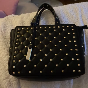 svart handväska  - säljer min snygga svarta handväska med guld nitar, den är köpt i lyserkyl i somras, den är knappt använd därför säljer jag den❤️. köpt för lite över 1000kr och säljs för 700kr, pris diskuterbart❤️.