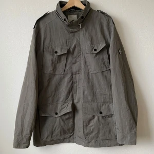 Grå Foreman jacka från J.Lindeberg - Säljer en stilren grå jacka från J.Lindeberg i storlek XL. Jackan har långa ärmar och flera praktiska fickor med knappar framtill. Perfekt för höst och vår med sin klassiska design och bekväma passform. Modell: Foreman 55 Space Nylon - Artnr: P194