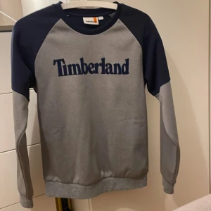 Grå och marinblå tröja från Timberland - Säljer en snygg grå och marinblå tröja från Timberland med deras logga på bröstet. Perfekt för en avslappnad stil. Tröjan är storlek 14 år. Listpris 935 kr.