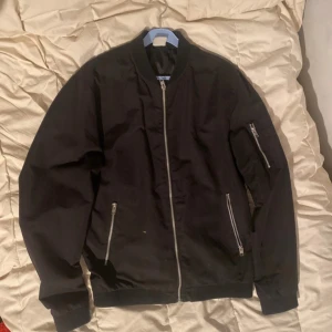 Svart bomberjacka från Jack & Jones - Snygg svart bomberjacka från Jack & Jones med dragkedja framtill och på ärmen. Jackan har ribbade muddar och två fickor med dragkedja. Perfekt för en stilren look.