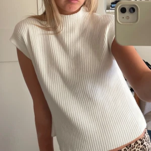 Vit ribbad topp - Säljer en stilren vit ribbad topp med hög hals och ärmlös design. Perfekt för en chic och minimalistisk look. Passar bra till både jeans och kjol.