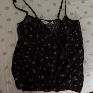 Svart blommig topp - Snygg svart linnetopp med små rosa blommor och justerbara axelband. Perfekt för varma sommardagar eller en avslappnad kvällslook.