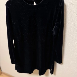 Svart sammetstunika från Lindex - Elegant svart tunika /klänning i mjukt sammetstyg från Lindex. Den har långa ärmar och en liten slitsdetalj. Perfekt för en stilren look.