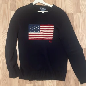Mörkblå tröja med amerikansk flagga från Polo Ralph Lauren - Säljer en snygg mörkblå tröja från Polo Ralph Lauren med en stickad amerikansk flagga på framsidan. Tröjan har långa ärmar och ribbade muddar vid ärmslut och nederkant. Perfekt för en stilren look! Köpt för 6000 i Ralph lauren butiken i stockholm. Kom privat om du vill ha bild på kvitto. Använd Max tre gånger, pris kan diskuteras! 
