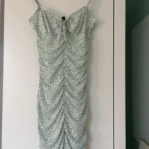 Blommig klänning från H&M - Söt blommig klänning från H&M i ljusgrön nyans med små blommor. Klänningen har smala axelband och en åtsittande passform med rynkade detaljer längs hela längden. Perfekt för sommarens varma dagar!