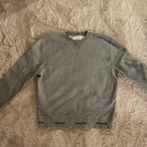Grå sweatshirt från Axel Arigato - Säljer en stilren grå sweatshirt från Axel Arigato. Tröjan har långa ärmar och en rund halsringning. Detaljer med 'Arigato' tryckt längs fållen och ärmsluten ger en unik touch. Knappt använd.
