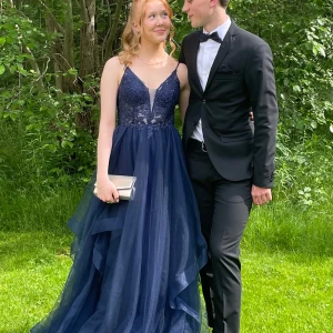 Mörkblå balklänning med spetsdetaljer - Mörkblå balklänning med spetsdetaljer och v-ringning. Klänningen har tunna axelband och en vacker öppen rygg.🥰 Kjolen är i flera lager av tyll. Klänningen är endast använd två gånger så är i fint skick. Axelbanden är uppsydda men kan läggas ner om man vill. Jag är 165 cm lång och hade låga klackar till och det funkade bra, men passar nog ännu bättre på någon som är lite längre. Färgen visas bäst på första bilden💙Stl 34. Ordinarie pris 3025 kr