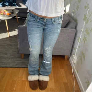 Blå jeans med slitningar - Säljer ett par blå jeans med slitningar på låren. De har en klassisk femficksdesign och är lågmidjade. Perfekta för en avslappnad stil.