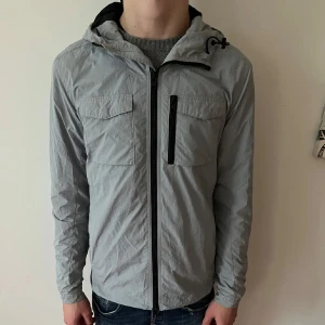 J Lindeberg windbreaker - Säljer en stilren grå vindjacka med huva och dragkedja. Jackan har två bröstfickor och en vertikal ficka med dragkedja. Perfekt för våren. Säljs inte längre då de är en äldre modell. Skriv gärna om ni undrar nåt! Pris kan diskuteras vid snabb affär 