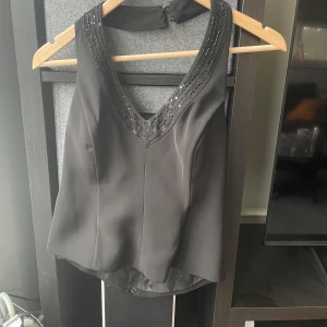 Svart ärmlös y2k topp med paljetter - Snygg svart ärmlös topp med paljetter längs den v-ringade halsringningen. Perfekt för en kväll ute eller en fest. En knappen är borta 