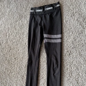Mörkgrå leggings från Stronger stXxs - Mörkgrå  leggings från Stronger med elastisk midja och textdetaljer. De har ett stilrent mönster med vita ränder på ena benet. Perfekta för träning eller en avslappnad dag.st Xxs