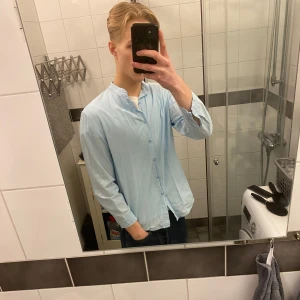 Ljusblå skjorta med ståkrage - Säljer en stilren ljusblå skjorta med ståkrage och knappar framtill. Skjortan har långa ärmar och en avslappnad passform, perfekt för en casual look. Passar bra till jeans eller chinos.