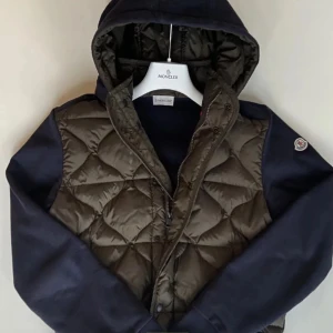 Quiltad jacka från Moncler - Snygg quiltad jacka från Moncler med huva och dragkedja. Jackan har en cool kombination av mörkblå ärmar och en olivgrön kropp. Perfekt för kyligare dagar med sin stilrena design och varma material. Nypris 9299 pris kan diskuteras vid snabb affär 