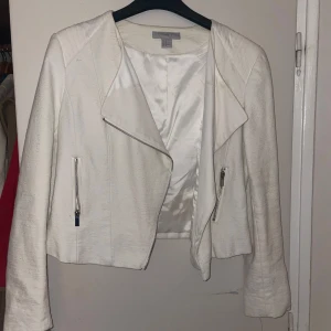 Vit jacka med dragkedjor - Snygg vit jacka med dragkedjor framtill och på fickorna. Jackan har en modern design med quiltade detaljer på axlarna och nedre delen av ryggen. Perfekt för att ge en stilren touch till din outfit.