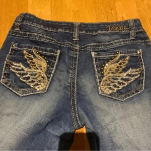 Blå jeans med broderade fickor - Snygga blå jeans från Vanity med unika broderade vingar på bakfickorna. Jeansen har en klassisk femficksdesign och en lätt slitning för en trendig look. Perfekta för dig som vill sticka ut med stil.