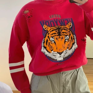 Röd sweatshirt med tigertryck - Cool röd sweatshirt med ett stort tigertryck och texten 'New York' på framsidan. Tröjan har långa ärmar med vita ränder och slitna detaljer för en edgy look. Perfekt för en avslappnad stil.