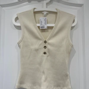 Beige ribbad linnetopp från H&M - Snygg beige ribbad linnetopp från H&M med v-ringning och dekorativa knappar framtill. Perfekt för en stilren och bekväm look. Passar bra till både jeans och kjol.