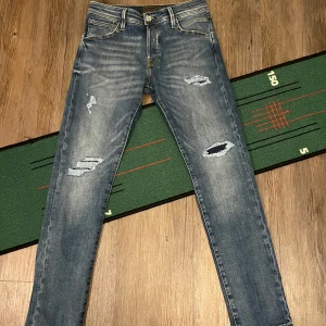 Blå jeans från Jack & Jones - Säljer ett par snygga blå jeans från Jack & Jones i modellen Slim/Glenn. De har en slim/regular passform och är dekorerade med slitningar för en trendig look. Perfekta för dig som gillar en stilren men avslappnad stil. Helt nya. W30 L30