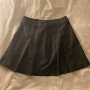 Svart kortkjol med veck - Säljer en snygg svart kortkjol med veck och knappdetalj. Perfekt för en stilren look. Kjolen har en klassisk design som passar till många olika outfits.