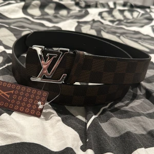 Louis Vuitton  - Säljer ett stilrent brunt bälte från Louis Vuitton med ett klassiskt rutigt mönster. Bältet har en iögonfallande LV-spänne i silverfärg. Perfekt för att ge din outfit en lyxig touch.