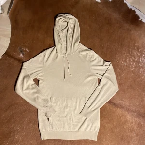 J Lindeberg hoodie - Hoodie i merino ull från j lindeberg, hoodien har en skön slim passform och skit mjukt och najs merino ull. Hoodien är i en unik beige färg och är perfekt nu till våren. Hoodien har endast en liten defekt på fickan men de märks inte när man har på sig den.