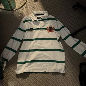 Vit och grön randig pikétröja från East West - Säljer en vit och grön randig pikétröja från East West med broderad emblem på bröstet. Tröjan har långa ärmar och en klassisk krage. Perfekt för en avslappnad stil med en touch av tradition.