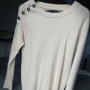 Beige Kashmir tröja med knappar - Säljer en stilren beige tröja i 100% Kashmir med dekorativa knappar på axeln. Tröjan har långa ärmar och en rund halsringning. Perfekt för en avslappnad och trendig look.