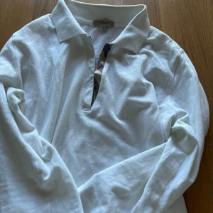 Vit pikétröja från Burberry - Säljer en stilren vit pikétröja från Burberry med klassisk krage och diskret logga på bröstet. Tröjan har långa ärmar och ett snyggt rutigt mönster på insidan av knappslån. Perfekt för en elegant och avslappnad look. Priset kan diskuteras.