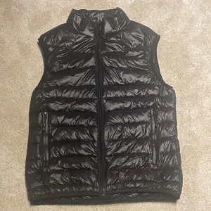 Svart dunväst från The North Face - Säljer en svart dunväst från The North Face. Västen har en dragkedja framtill och två praktiska fickor med dragkedjor. Perfekt för kyligare dagar när du vill hålla värmen utan att bära en jacka.