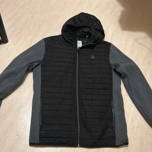 Jack & Jones cardigan - Säljerw en stilren svart och grå jacka från Jack & Jones. Jackan har en quiltad design med dragkedja och huva. Perfekt för kyligare dagar med sin långärmade stil och praktiska fickor.