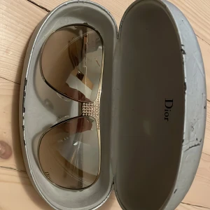 Solglasögon från Dior med strassdetaljer - Snygga solglasögon från Dior med bruna tonade glas och guldfärgad ram. Glasögonen har en elegant strassdetalj på sidorna som ger en lyxig känsla. Perfekt accessoar för soliga dagar. Kommer med etui. TA KONTAKT OM TARDES OG PRIS. TAR TRADES.