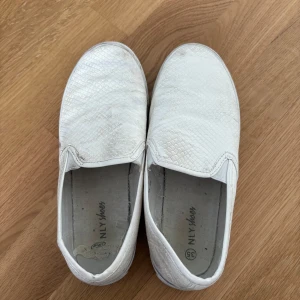 Vita slip-on skor från NLY Shoes - Säljer ett par vita slip-on skor från NLY Shoes med ett diskret ormskinnsmönster. Skorna har en bekväm passform och är perfekta för en avslappnad stil. De är lätta att ta på och av.