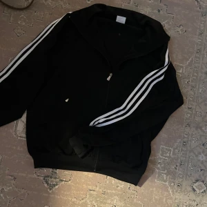 Svart träningsjacka från Adidas - Säljer en svart träningsjacka från Adidas med klassiska vita ränder längs ärmarna. Jackan har dragkedja framtill och är långärmad. Perfekt för sportiga aktiviteter eller en avslappnad stil.