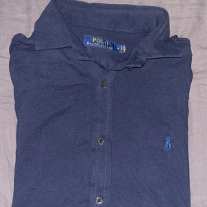 Mörkblå pikétröja från Ralph Lauren - Säljer en klassisk mörkblå pikétröja från Ralph Lauren med den ikoniska broderade loggan på bröstet. Tröjan har knappar framtill och en stilren krage. Perfekt för en avslappnad men ändå stilfull look.