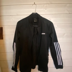 Svart träningsjacka från Adidas - Säljer en svart träningsjacka från Adidas med klassiska vita ränder längs ärmarna. Jackan har en dragkedja framtill och Adidas-loggan på bröstet. Perfekt för sport och träning.