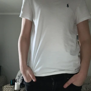 Vit t-shirt från Ralph Lauren - Säljer en klassisk vit t-shirt från Ralph Lauren med det ikoniska broderade logotypen i blått på bröstet. T-shirten har en slim passform och är tillverkad i mjuk bomull. Perfekt för en stilren look.