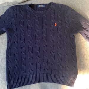 Mörkblå stickad tröja från Ralph Lauren - Säljer en snygg mörkblå stickad tröja från Ralph Lauren med klassiskt kabelstickat mönster. Tröjan har en liten  logga på bröstet och ribbade kanter vid ärmslut och nederkant. Perfekt för kyliga dagar!