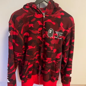 Röd och svart camo hoodie från A Bathing Ape x FaZe Clan - Säljer en cool röd och svart camo hoodie från A Bathing Ape i samarbete med FaZe Clan. Hoodien har dragkedja och en stor logga på ryggen. Perfekt för dig som gillar streetwear och gaming.