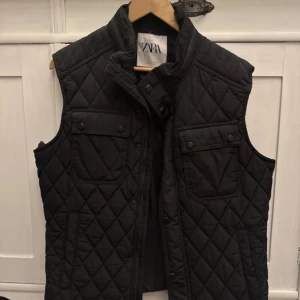 Svart quiltad väst från Zara - Säljer en stilren svart quiltad väst från Zara. Västen har knappar framtill och två bröstfickor med knappstängning. Perfekt för lager-på-lager under kyligare dagar.