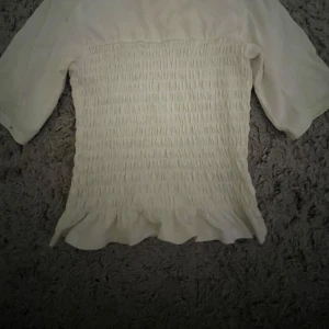 Vit smockad topp - Säljer en söt vit topp med smockad design och volangdetaljer i nederkant. Toppen har trekvartsärmar och en bekväm passform. Perfekt för en stilren look! Köpt i somras men använd få gånger därav bra skick! Säljs ej längre!!!😊