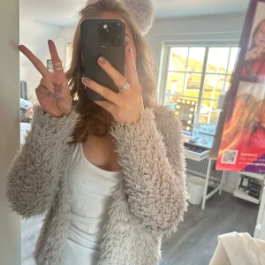 Grå teddyjacka från Gina Tricot - Säljer en supermjuk och fluffig grå teddyjacka från Gina Tricot. Jackan har en öppen design utan knappar eller dragkedja och är perfekt för att hålla sig varm och stilren. Den är långärmad och har en härlig textur som ger en mysig känsla.